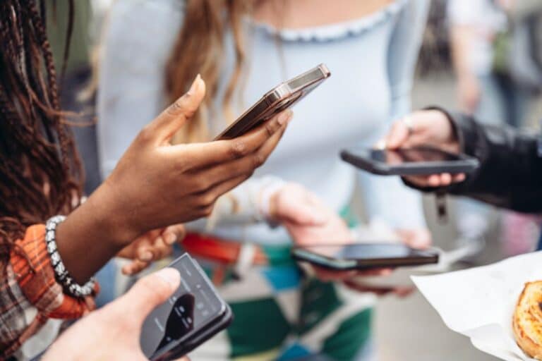 A group of people standing close together, each holding and using their mobile phones online. Their faces are not visible, with the focus on their hands and devices.