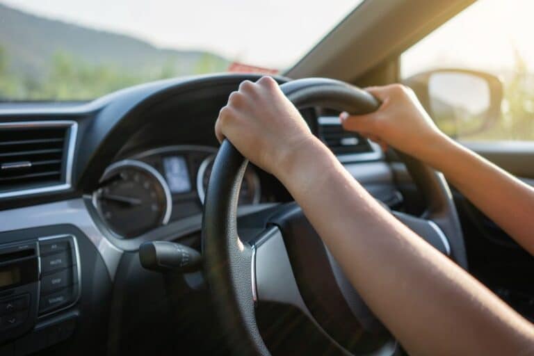 Close-up of a person’s hands gripping a steering wheel while sharing a car, sunlight streaming through the window and the dashboard visible in the background.
