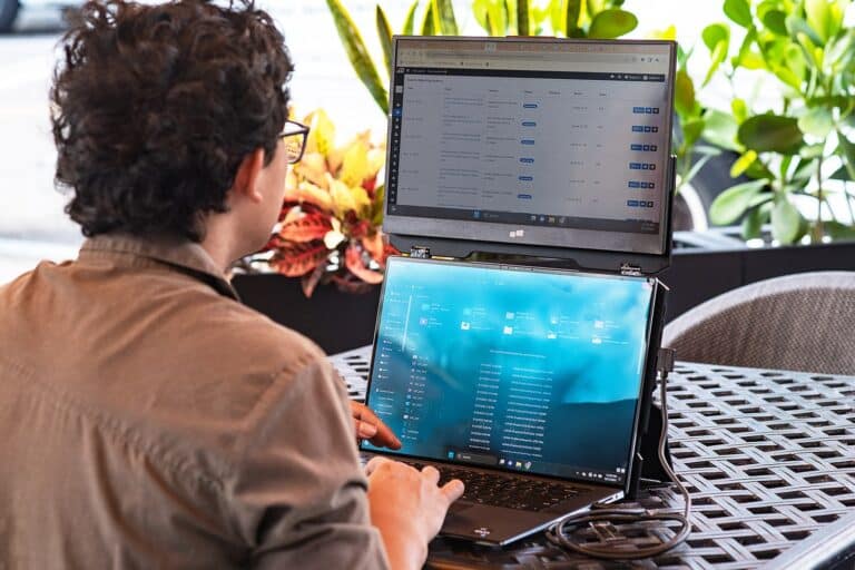A person with curly hair uses a dual-screen laptop setup with an extra screen stacked above the main one at an outdoor table, surrounded by green plants in the background.