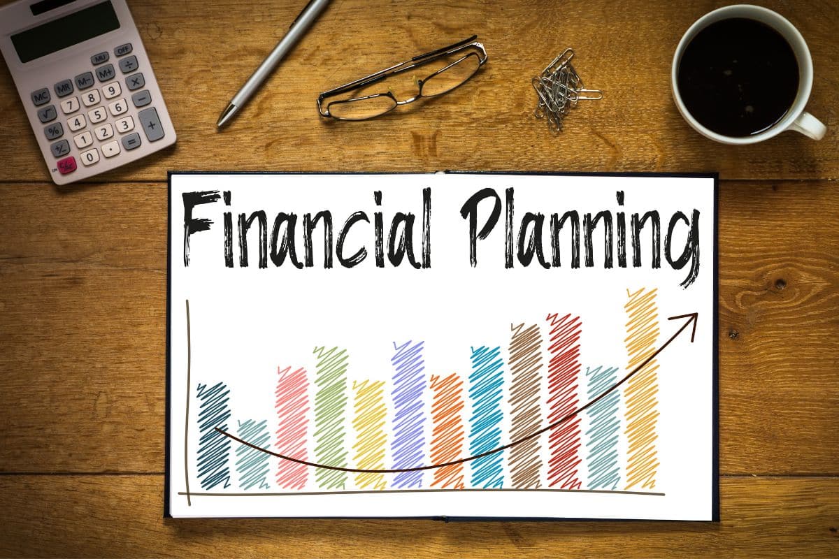 A desk with a notebook displaying "Financial Planning" above a colourful bar graph and upward arrow, surrounded by a calculator, pen, glasses, paper clips, and a cup of coffee—tools to help reduce financial stress.