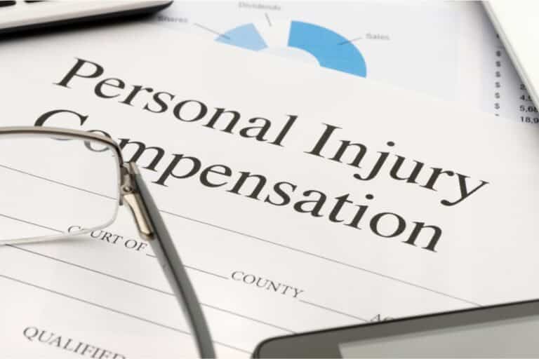 A document titled "Personal Injury Compensation" sits on a desk, partially covered by spectacles and a pen, with financial charts and personal injury paperwork visible in the background.