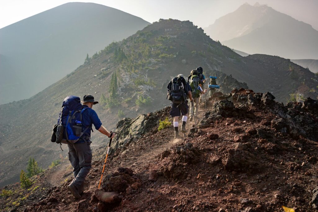 A group of hikers with rucksacks embark on an adventure, trekking along a rugged, rocky mountain ridge under a hazy sky, surrounded by sparse greenery and distant peaks.