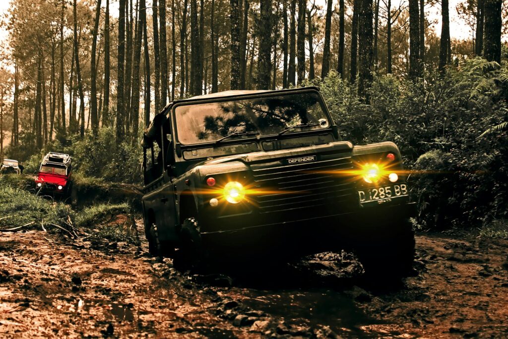 A black off-road vehicle with headlights on drives through a muddy forest trail, embodying the spirit of adventure, whilst another vehicle follows in the background amongst tall trees and dense greenery.