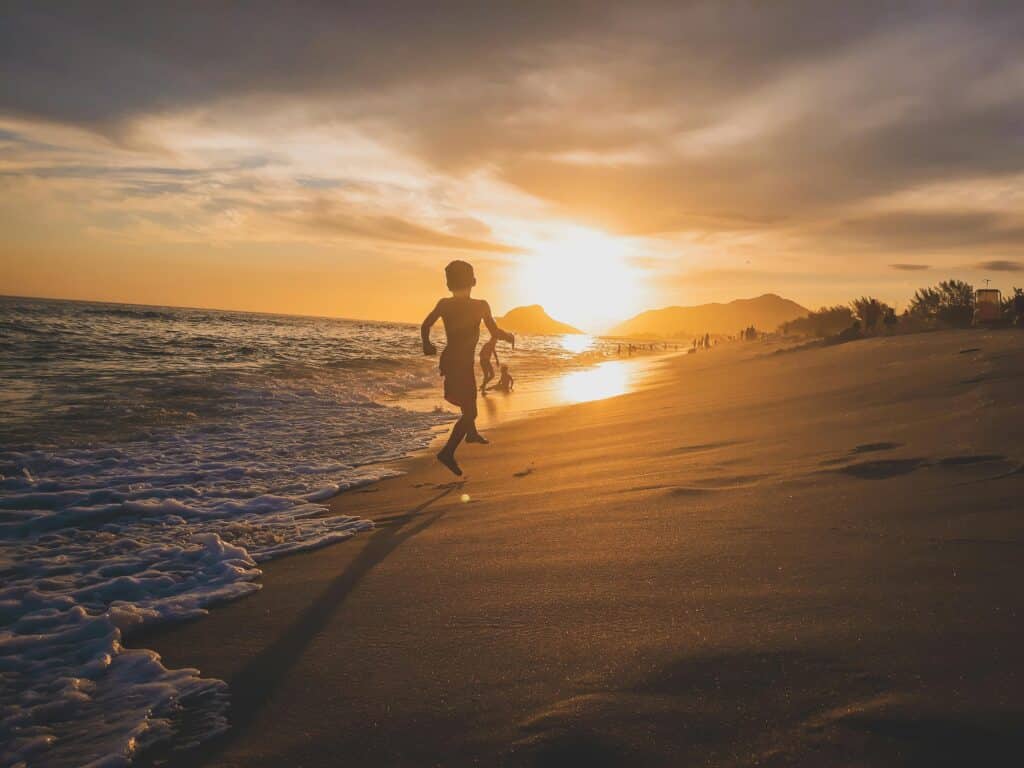 A child runs along the shoreline at sunset, embracing adventure as gentle waves wash onto the sandy beach. The sky is vibrant with orange and yellow hues, mountains are visible in the distance, and other people walk in the background.