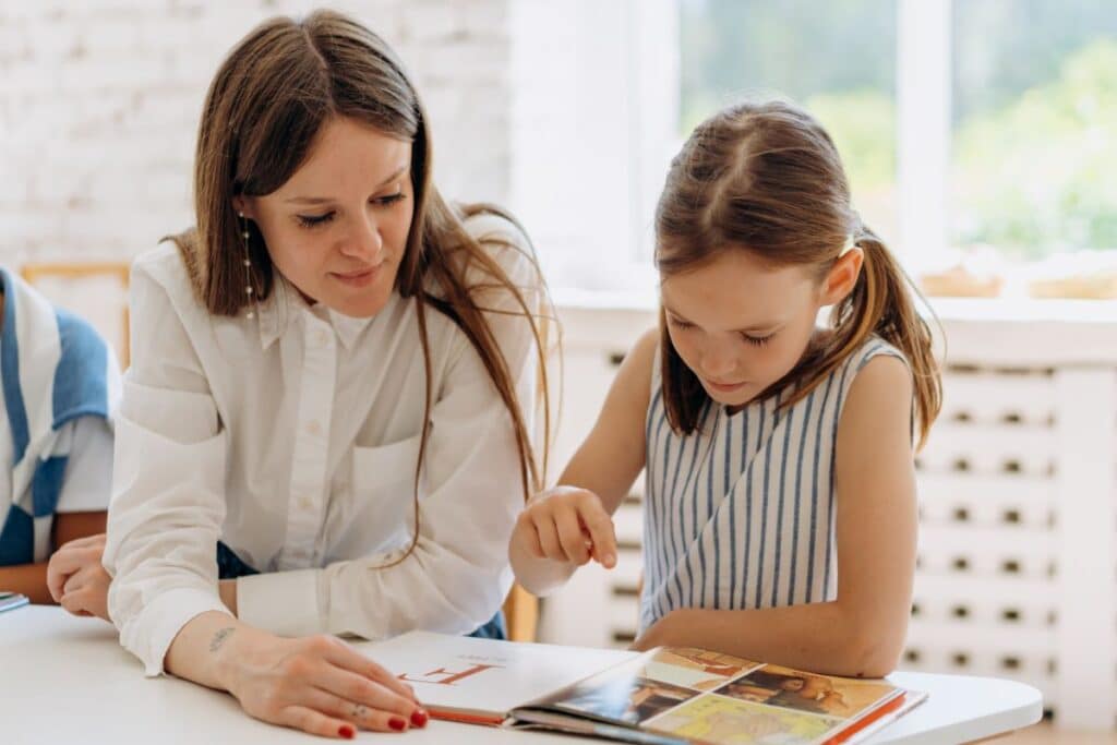 A woman sits beside a young girl who is reading a book and pointing at a page, as they build literacy skills together. The woman watches attentively, and they are seated at a bright table in a well-lit room.