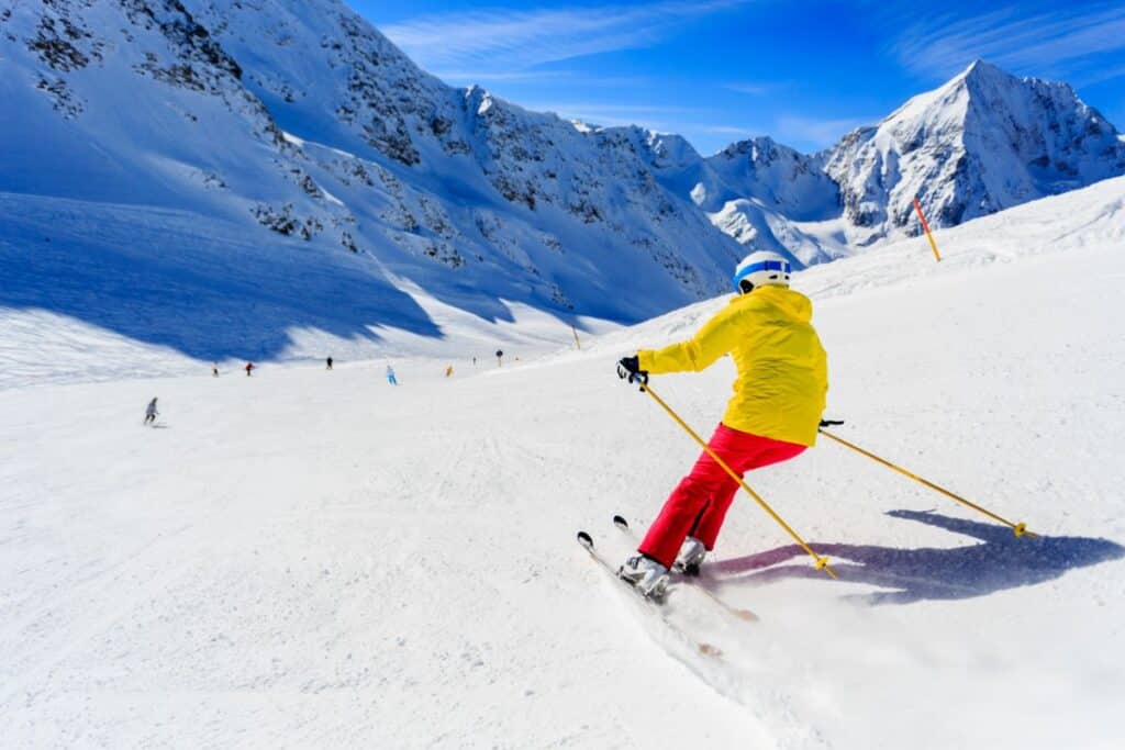 A skier in a yellow jacket and red trousers enjoys skiing downhill on a snowy mountain slope under a bright blue sky, with other skiers and snow-covered peaks visible in the background.
