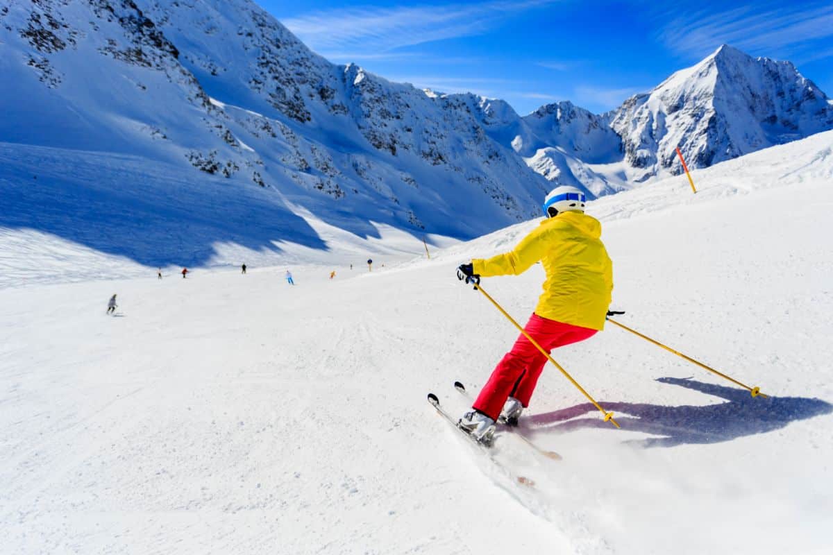 A skier in a yellow jacket and red trousers enjoys skiing downhill on a snowy mountain slope under a bright blue sky, with other skiers and snow-covered peaks visible in the background.