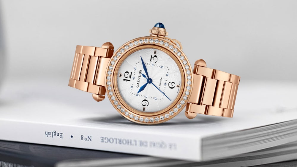 A luxurious gold wristwatch from the Cartier Ladies' Watches collection, with a diamond-studded bezel and blue crown, lies on a closed book, showcasing its white face, blue hands, and elegant Arabic numerals.