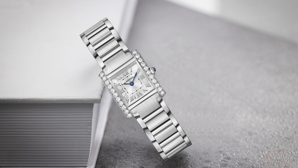 A silver luxury Cartier Ladies' Watch with a rectangular face, diamond-encrusted bezel, Roman numerals, and blue hands rests elegantly against the edge of a closed white book on a grey surface.