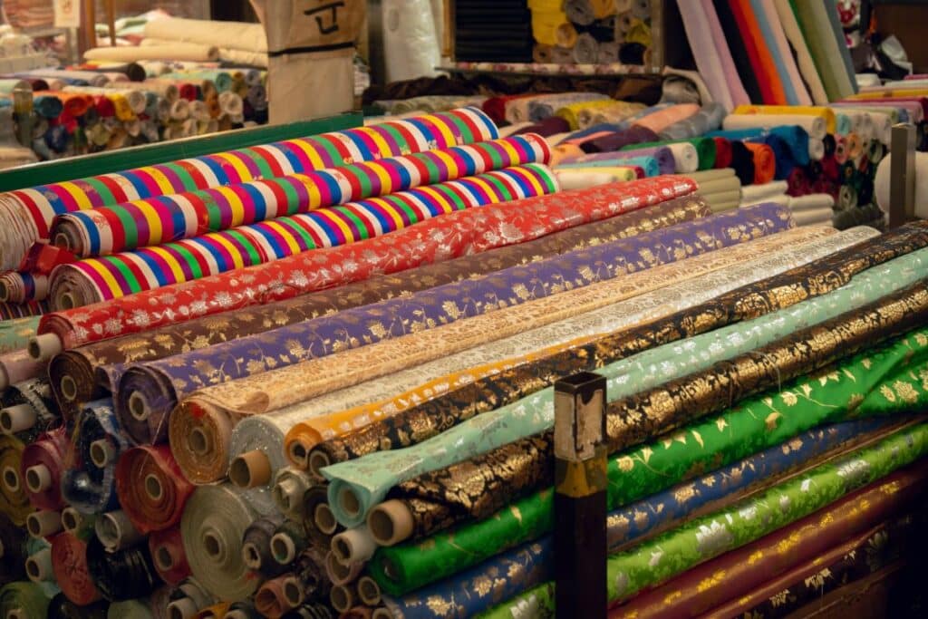 Several rolls of colourful fabric with various patterns, including floral designs and metallic accents, are stacked on display in a fabric shop. Some fabrics showcase bold stripes, while others feature intricate fabric motifs.