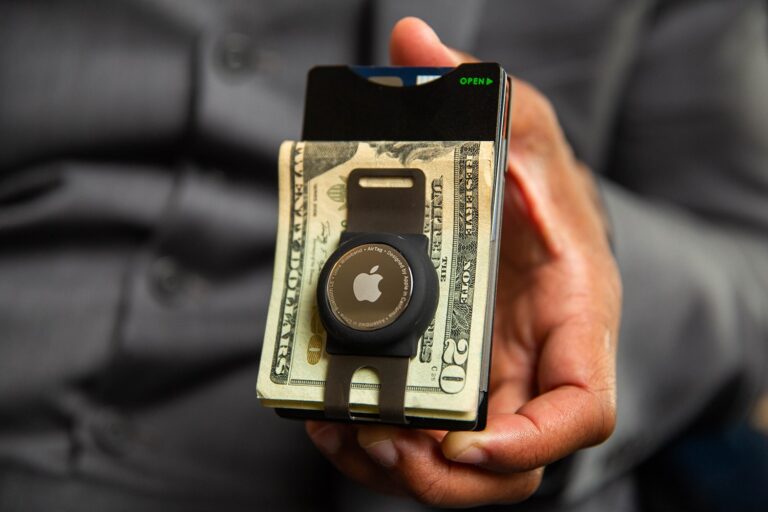 A hand holds a minimalist wallet money clip with folded cash, credit cards, and an Apple AirTag attached, all set against the background of a grey shirt.