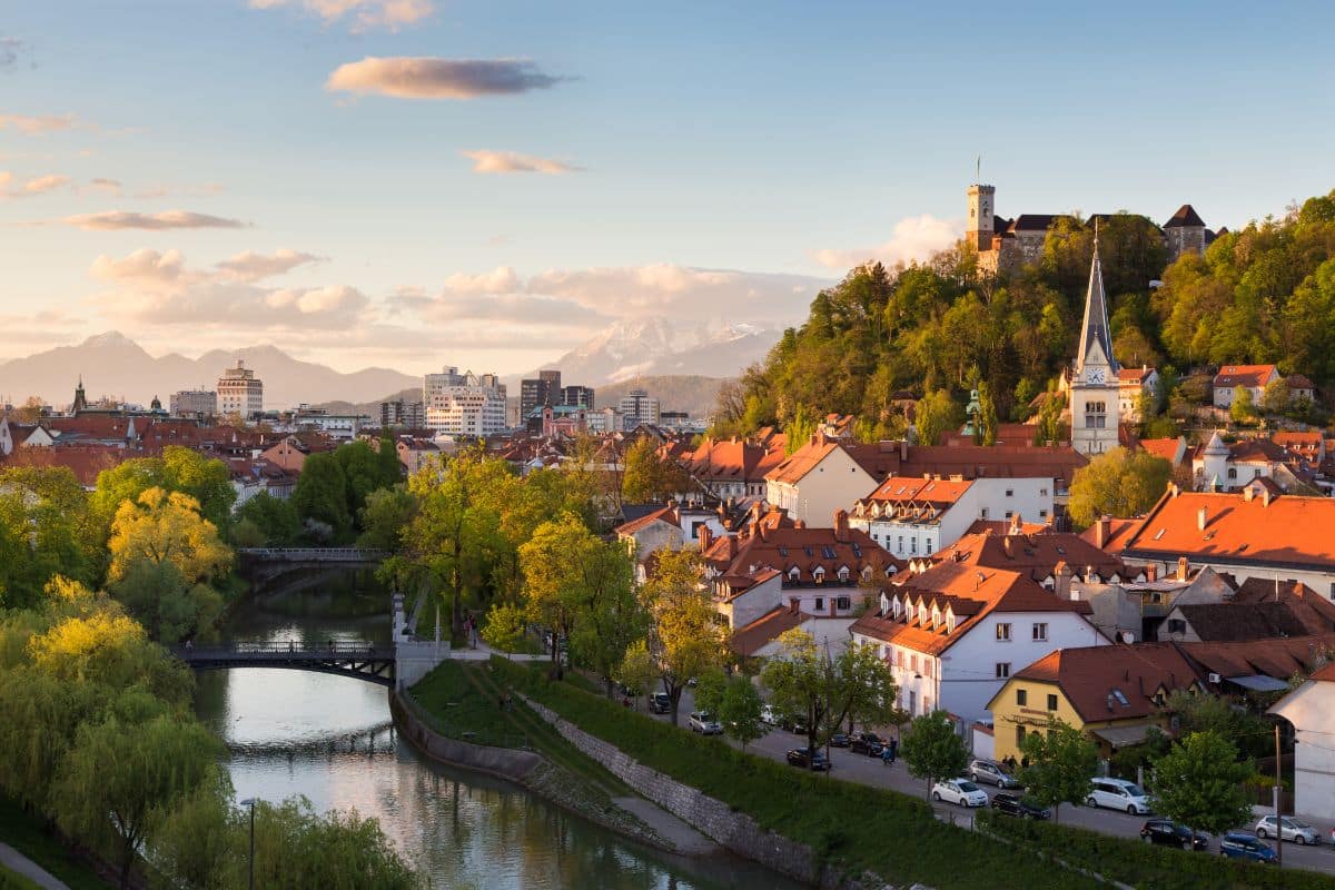 A scenic holiday view of Ljubljana, Slovenia, with red-roofed buildings, a river winding through the city, green trees, and Ljubljana Castle perched atop a hill under a partly cloudy sky at sunset.