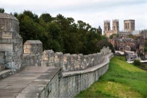 A stone city wall curves through green grass and trees, inviting scenic walks past historic buildings and the towers of a large cathedral rising in the background under a cloudy sky.