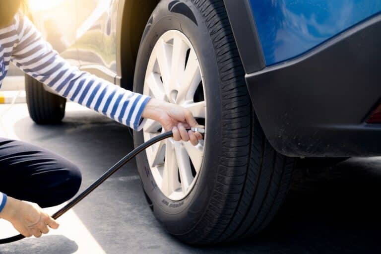 A person in a striped shirt kneels beside a blue car, inflating its tyre with an air hose at a petrol station under the bright sunlight.