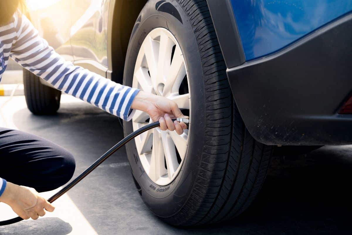 A person in a striped shirt kneels beside a blue car, inflating its tyre with an air hose at a petrol station under the bright sunlight.