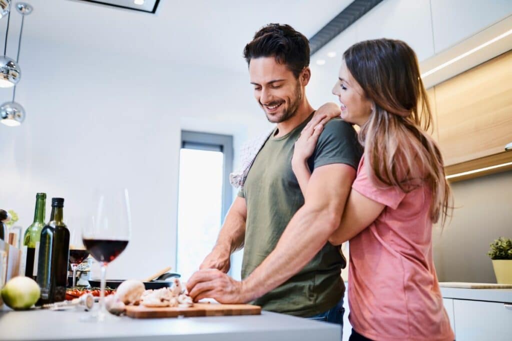 A smiling man chops vegetables on a kitchen worktop while a woman hugs his shoulder, both enjoying a cosy date night. Bottles, a glass of red wine, and chopped ingredients are spread out on the worktop.