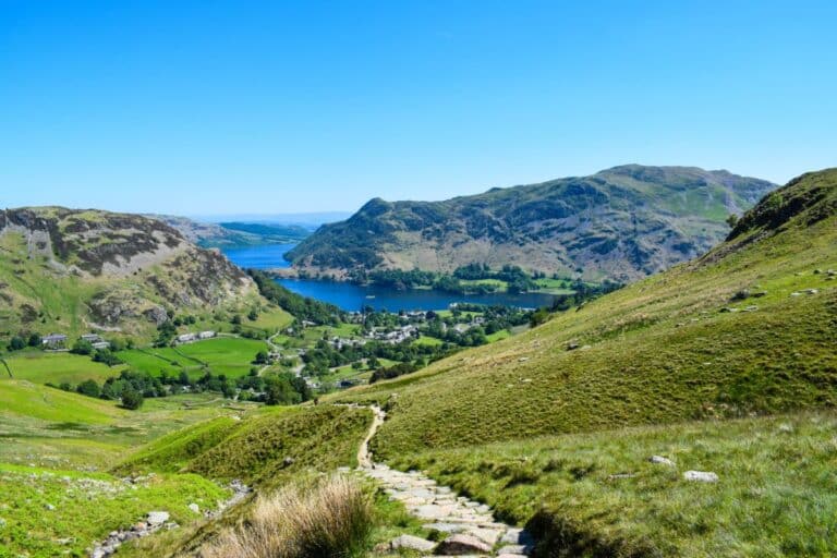 A winding stone path leads down a grassy hillside towards a village by a blue lake, surrounded by green fields and rolling hills—scenery reminiscent of national parks—under a clear blue sky.