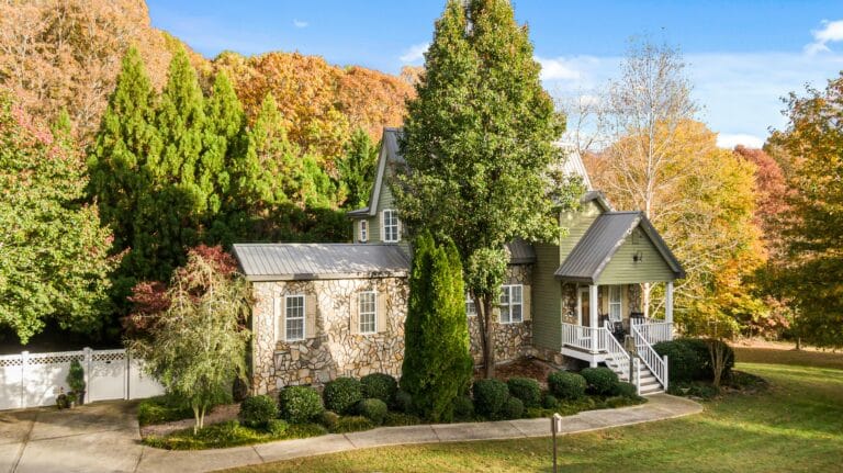 A stone and wood house with a front porch boasts impressive kerb appeal, surrounded by green shrubs and tall trees, set against a backdrop of autumn foliage under a clear blue sky.