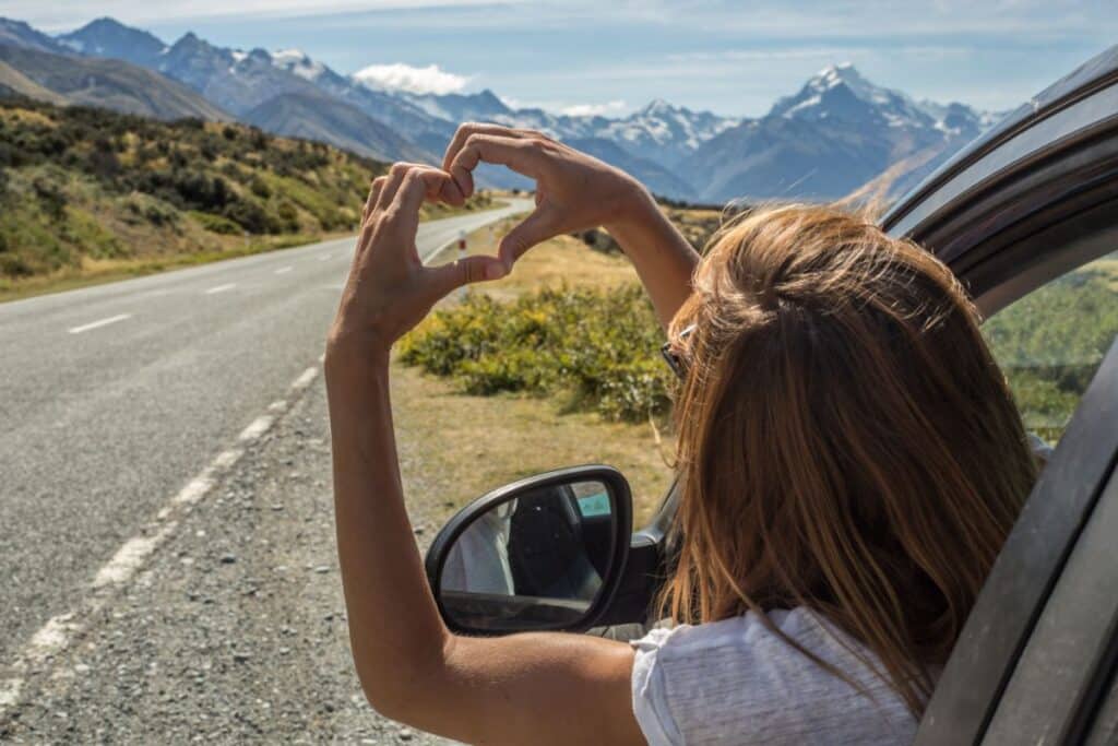 A person leans out of a car window on a scenic mountain road trip, making a heart shape with their hands. Snow-capped peaks and blue sky are visible in the background.