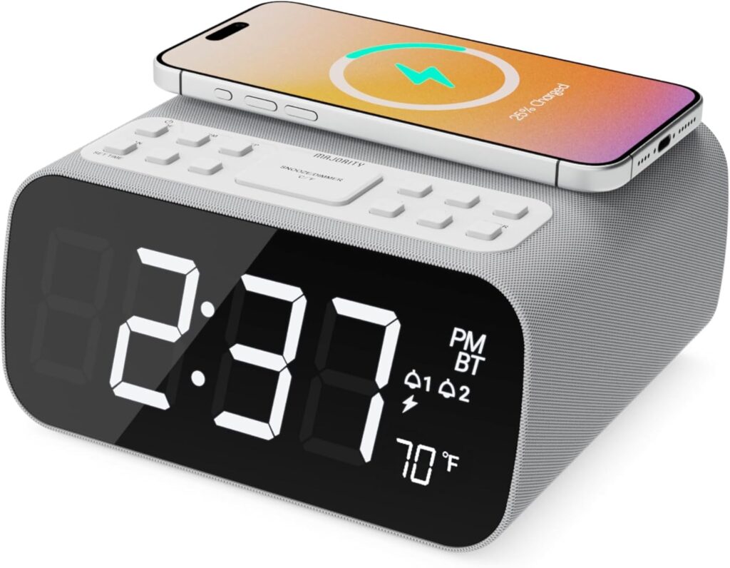 A digital alarm clock with a large LED display showing 2:37 pm and 21°C, featuring various buttons on top and a smartphone wirelessly charging—a perfect Father’s Day gift for organised mornings.