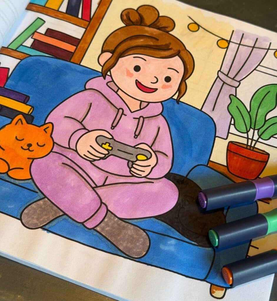 A colouring book page shows a smiling person in a pink hoodie playing video games on a blue sofa, with an orange cat beside them. There are bookshelves, a plant, and fairy lights in the background. Four coloured markers are in the corner.