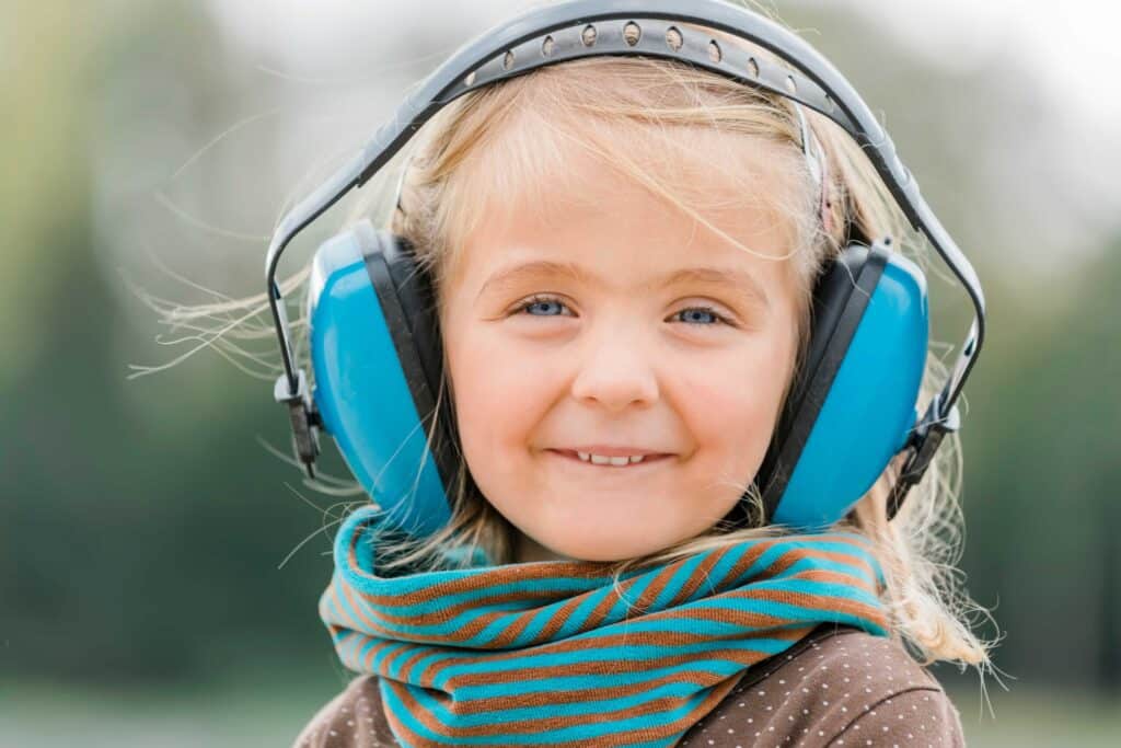 Smiling young girl with blonde hair wearing large blue noise-cancelling headphones for better hearing and a striped scarf, standing outdoors with a blurred green background.