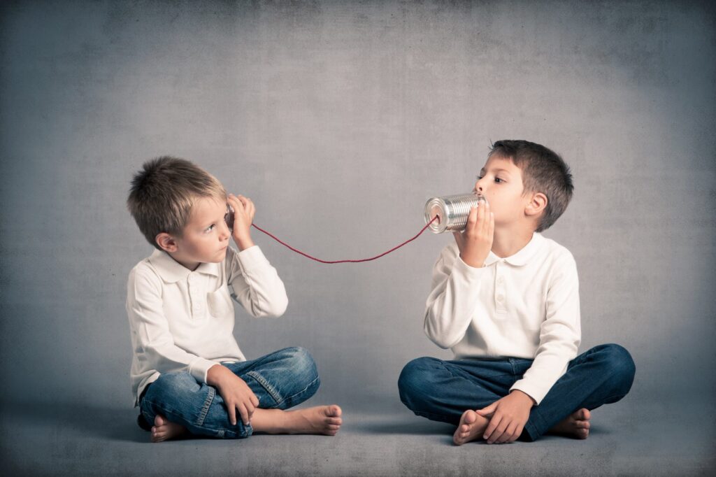 Two young boys sit cross-legged on the floor, facing each other. One boy speaks into a tin can connected by a string, while the other holds it to his ear, listening closely and enjoying the simple joy of hearing through their homemade telephone.