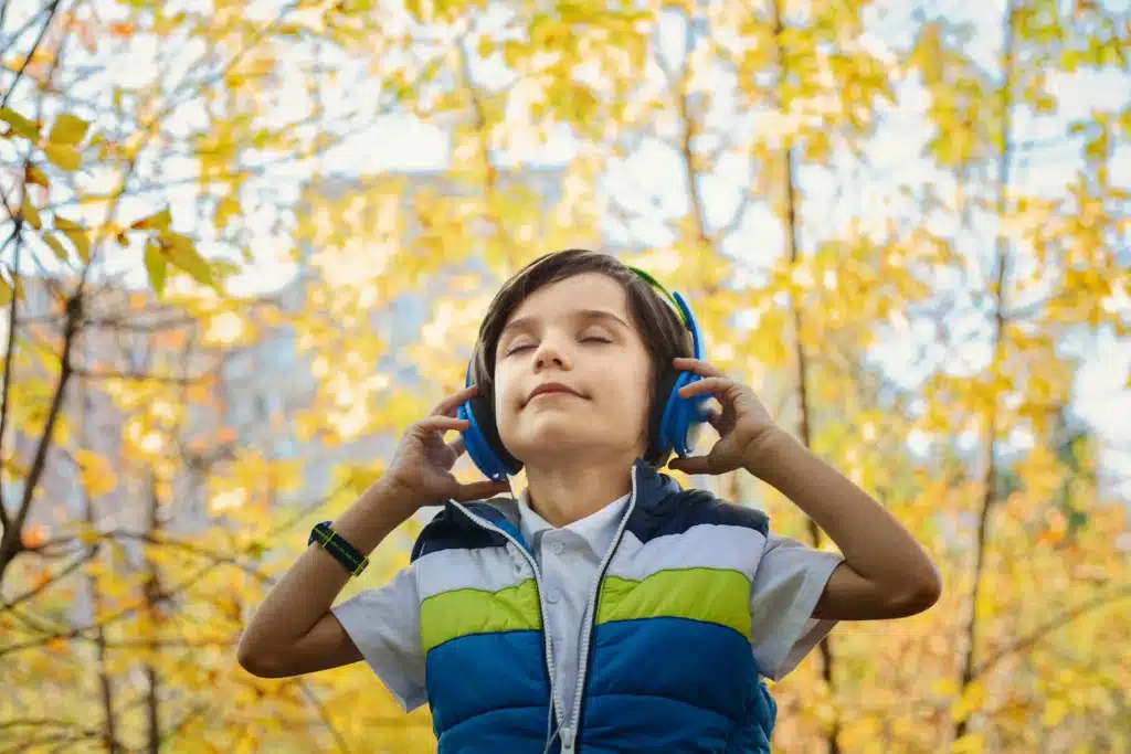 A young boy wearing a blue waistcoat and headphones stands outdoors among autumn trees, eyes closed and smiling, hearing music on a sunny day.