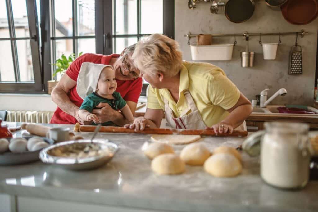 An elderly couple smiles and leans over a kitchen counter with their grandchild, who wears a chef’s hat. Baking together, they share joyful moments and hobbies, surrounded by dough, eggs, and ingredients on the sunlit worktop.