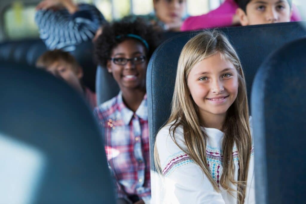 A group of children enjoy transport on a school bus. In the foreground, a smiling blonde girl in a white shirt faces the camera, whilst other kids sit behind her, including a girl in glasses and a checked shirt.