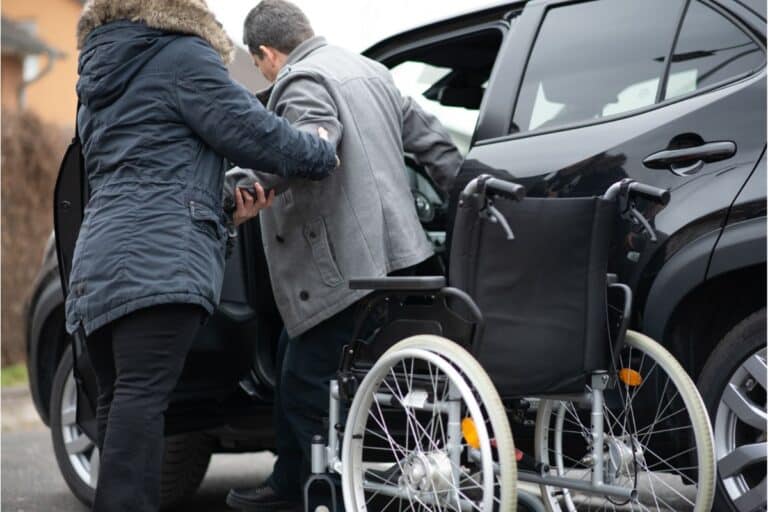 A person assists a man in a grey coat with family transport, helping him transfer from a wheelchair into a black car, while the empty wheelchair rests on the pavement nearby.