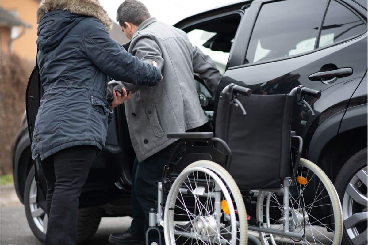 A person assists a man in a grey coat with family transport, helping him transfer from a wheelchair into a black car, while the empty wheelchair rests on the pavement nearby.