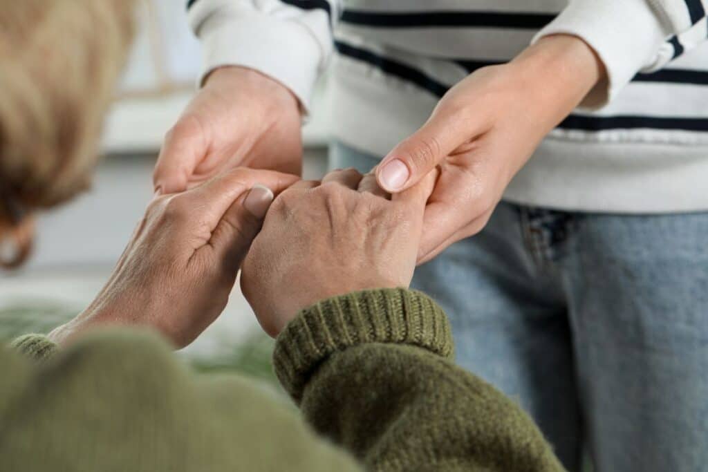 A person wearing a striped shirt gently holds the hands of an older individual in a green jumper, conveying comfort and support in a warm home setting.