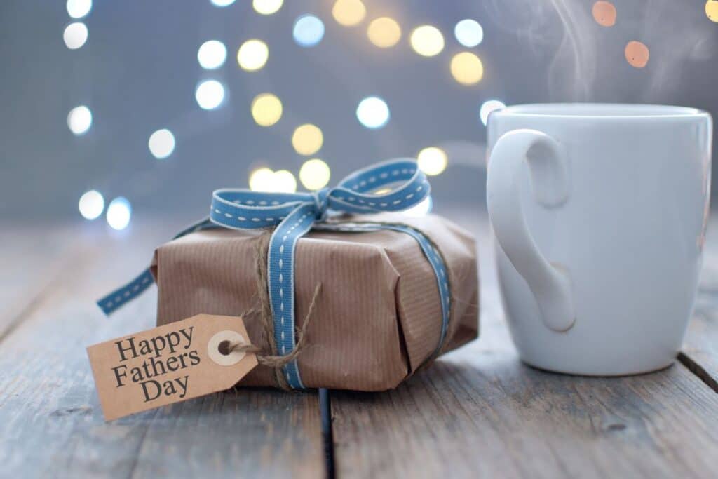 A wrapped present with a blue ribbon and a "Happy Father's Day" tag sits next to a steaming white mug on a wooden table, capturing the warm spirit of Father's Day amid softly blurred lights in the background.