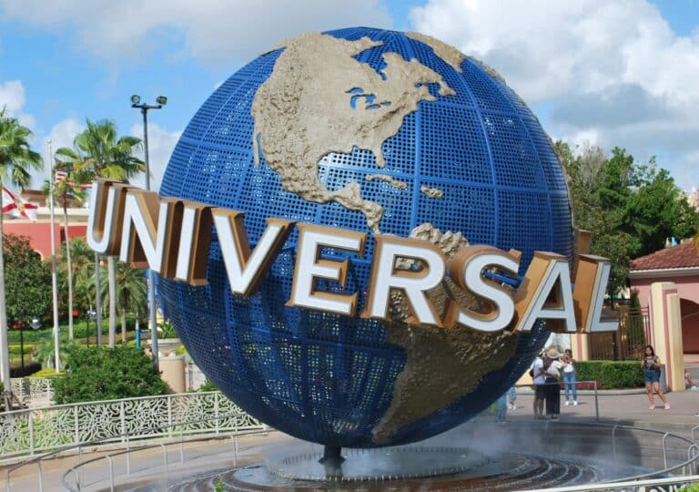 A large blue globe with gold continents and the word "UNIVERSAL" in bold white letters stands as an entrance monument at Universal Studios, offering a distinct contrast to Disney, surrounded by palm trees and visitors.