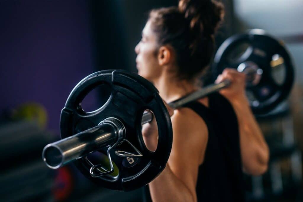 A woman with dark hair in a bun is powering through her workout, lifting a barbell loaded with weights across her shoulders in a gym. The barbell takes centre stage, while she remains slightly out of focus in the background.
