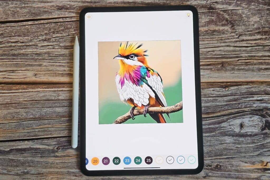 A tablet perfect for iPad colouring displays a vibrant digital drawing of a bird with orange, yellow, purple, green, and white feathers perched on a branch. A stylus rests beside the tablet on a wooden surface.