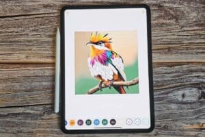 A tablet perfect for iPad colouring displays a vibrant digital drawing of a bird with orange, yellow, purple, green, and white feathers perched on a branch. A stylus rests beside the tablet on a wooden surface.
