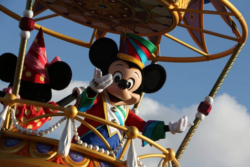 Mickey Mouse, dressed in a colourful costume, waves from a decorated Disney parade float alongside another character in a red outfit. The sky is blue with some clouds in the background.