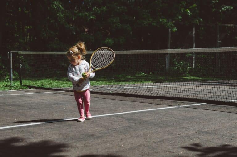 A young girl in pink trousers and a jumper stands on an outdoor tennis court, holding a tennis racket and ball, preparing to play—a joyful moment in a child’s life. The net and lush green trees are visible in the background.