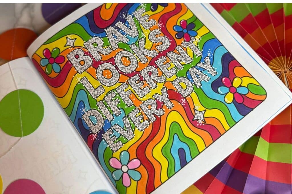 A colourful colouring book page with rainbow waves, flowers, and stars, featuring the glittery text “BRAVE LOOKS DIFFERENT EVERY DAY.” Bright decorations and a colourful paper fan are partly visible around the page.