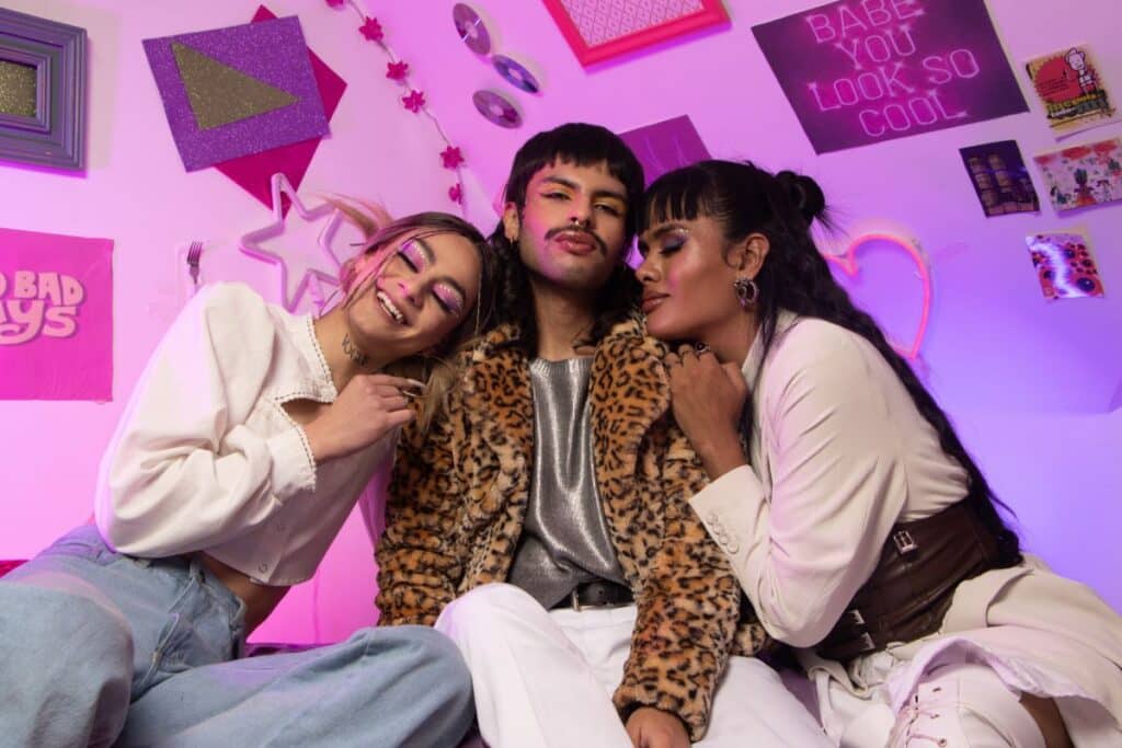Three people pose happily together in a colourful, Y2K-inspired retro room with pink and purple lighting. They wear stylish, bold outfits and make-up. Poster on the wall reads "BABE YOU LOOK SO COOL.