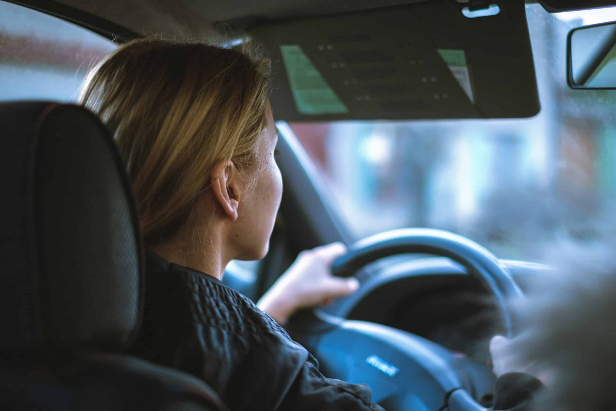 A person with blonde hair drives a car, holding the steering wheel with both hands and looking through the windscreen, ensuring they're safe on the road. The photo is taken from the back seat, focusing on the driver's side.