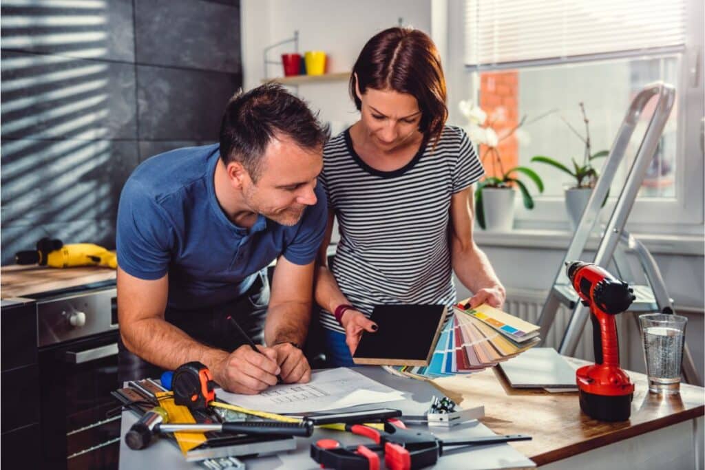 A man and woman review papers, colour swatches, and an insulation board at a table filled with tools and a cordless drill, planning a home renovation in a bright, modern kitchen.