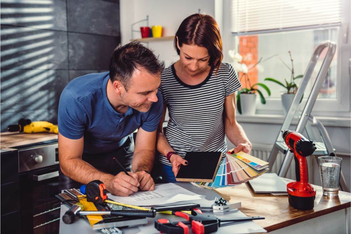 A man and woman review papers, colour swatches, and an insulation board at a table filled with tools and a cordless drill, planning a home renovation in a bright, modern kitchen.