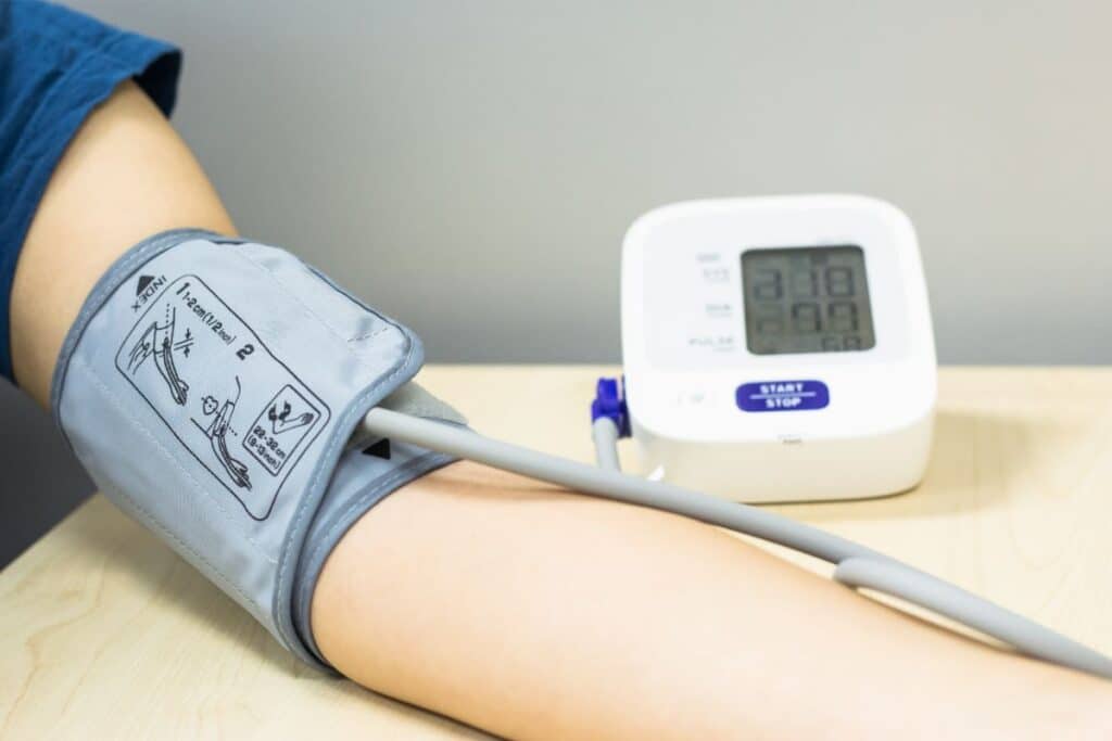 A person’s arm with a blood pressure cuff wrapped around it, connected by a tube to an electronic blood pressure monitor on a wooden table—an essential step in tracking healthy blood pressure. The monitor display is switched on but not fully legible.