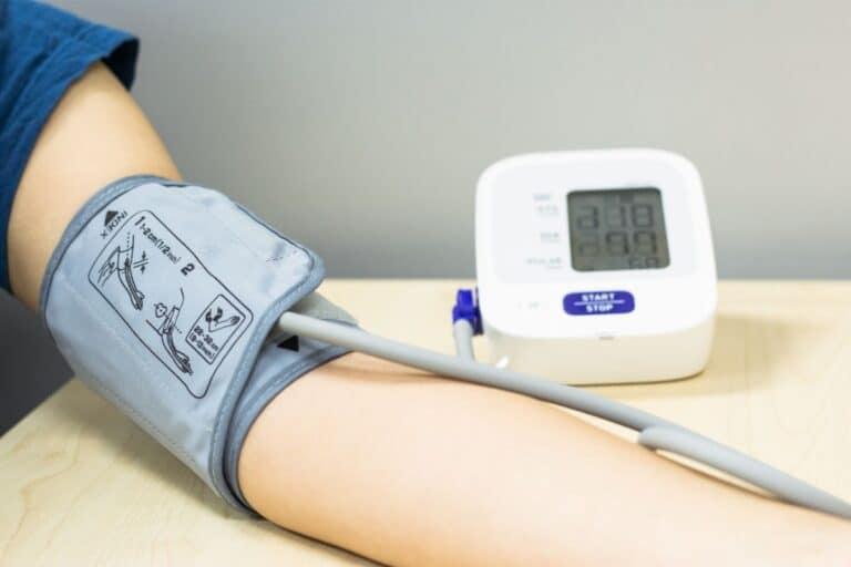 A person’s arm with a blood pressure cuff wrapped around it, connected by a tube to an electronic blood pressure monitor on a wooden table—an essential step in tracking healthy blood pressure. The monitor display is switched on but not fully legible.