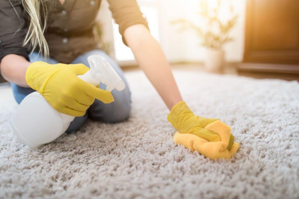 A person wearing yellow rubber gloves works to remove ink from carpets, cleaning a light-coloured carpet with a yellow cloth and spraying solution from a white bottle as sunlight shines in the background.