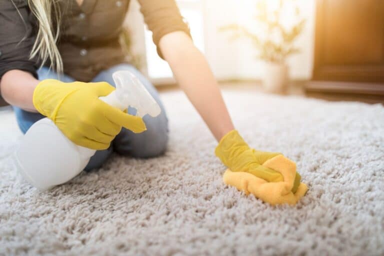 A person wearing yellow rubber gloves works to remove ink from carpets, cleaning a light-coloured carpet with a yellow cloth and spraying solution from a white bottle as sunlight shines in the background.