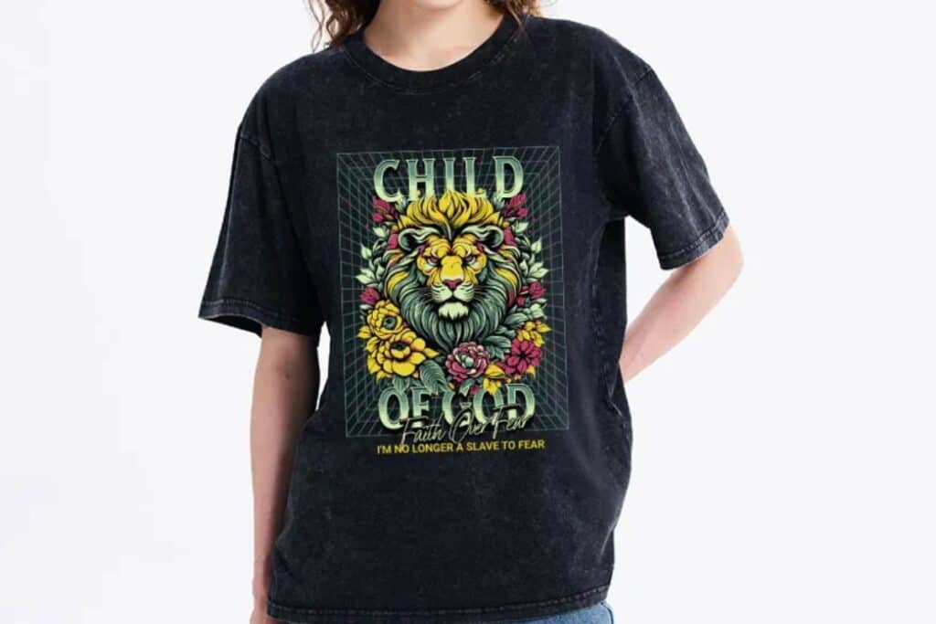 A person wearing a black Christian clothing T-shirt featuring a colourful lion and floral graphic. The shirt reads "CHILD OF GOD" and "I’m No Longer A Slave To Fear" in bold letters.